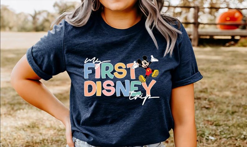 May include: Navy blue t-shirt with the text "My First Disney Trip" in colorful letters and a graphic of Mickey Mouse. The shirt is a casual style, suitable for a Disney vacation or themed event.