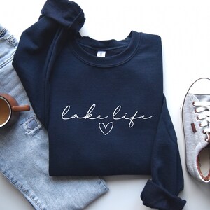 K&ouml;nnte beinhalten: Ein marineblauer Pullover mit dem Schriftzug "lake life" in wei&szlig;er Schrift mit einem kleinen Herz darunter. Der Pullover liegt auf einer wei&szlig;en Oberfl&auml;che mit einem Paar grauen Canvas-Schuhen und einer blauen Jeans.
