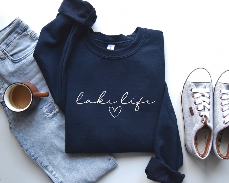 K&ouml;nnte beinhalten: Ein marineblauer Pullover mit dem Schriftzug "lake life" in wei&szlig;er Schrift mit einem kleinen Herz darunter. Der Pullover liegt auf einer wei&szlig;en Oberfl&auml;che mit einem Paar grauen Canvas-Schuhen und einer blauen Jeans.