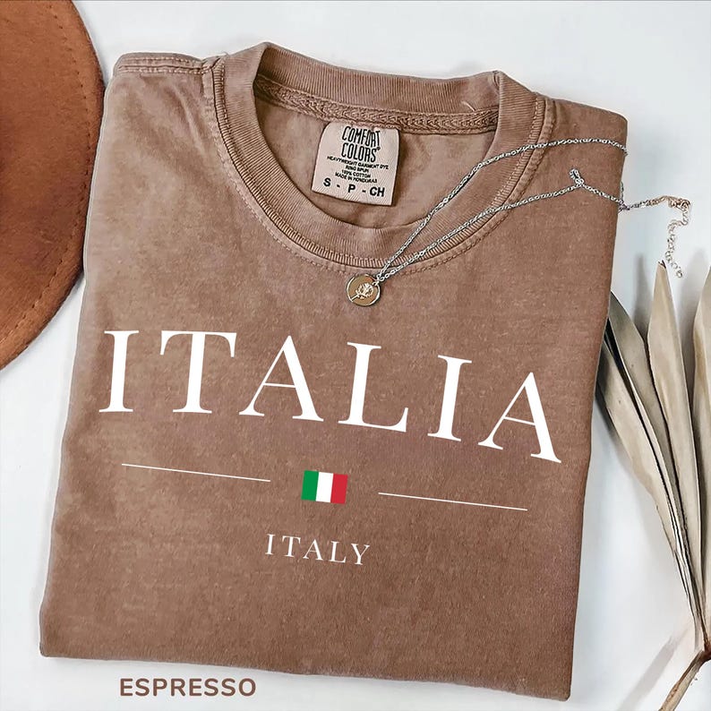 May include: Espresso-colored t-shirt with the word "ITALIA" in white, a line, and the Italian flag. Below, the word "ITALY" is printed. A silver necklace with a pendant rests on the shirt. The shirt is folded.