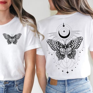 May include: Two white t-shirts featuring a black butterfly graphic. One shirt has a butterfly on the front and the other has a butterfly with a crescent moon and stars on the back.