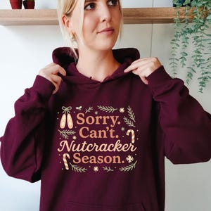 May include: Burgundy hoodie with the text "Sorry. Can't. Nutcracker Season." The text is in a festive font with decorative elements like ballet shoes, candy canes, and stars. The hoodie has a hood and a relaxed fit.