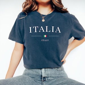 May include: A navy blue t-shirt with the word "ITALIA" in white capital letters. Below the word is a horizontal line, a small Italian flag, and the word "ITALY". The shirt is a casual style.