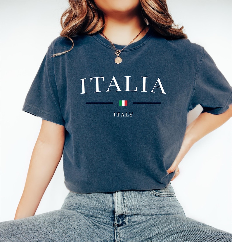 May include: A navy blue t-shirt with the word "ITALIA" in white capital letters. Below the word is a horizontal line, a small Italian flag, and the word "ITALY". The shirt is a casual style.