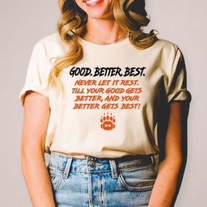 May include: Cream-colored t-shirt with the text "GOOD. BETTER. BEST. NEVER LET IT REST. TILL YOUR GOOD GETS BETTER, AND YOUR BETTER GETS BEST!" in black and orange. A bear paw graphic with the word "WIN" is below the text.