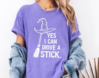 May include: A purple t-shirt with the text "YES I CAN DRIVE A STICK." The graphic features a witch's hat and broom in white. The shirt has short sleeves and a relaxed fit, perfect for Halloween.