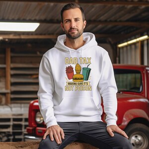 May include: White hoodie featuring the text "DAD TAX" and a graphic of fries, a burger, ice cream, and a drink. The text "MAKING SURE IT'S NOT POISON" is below the graphic. The hoodie is worn by a person.