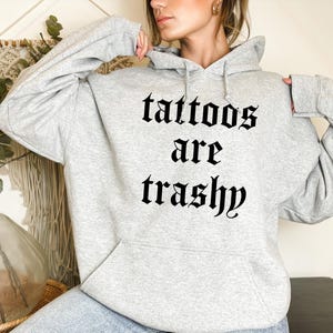 May include: Gray hooded sweatshirt with the text "tattoos are trashy" printed in black gothic font.