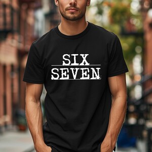May include: Black crew neck t-shirt with the white text "SIX SEVEN" printed across the chest. The text is separated by a horizontal line. The shirt is made of a soft material.