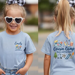 Pu&ograve; includere: Una t-shirt azzurra con una piccola grafica sul petto sinistro con il nome Emily e il testo Cousin Camp 2026. Il retro mostra una grafica pi&ugrave; grande con il testo Cousin Camp Mama and Papa 2026 e varie icone colorate.