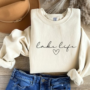 K&ouml;nnte beinhalten: Ein cremefarbener Pullover mit dem Text "lake life" und einem kleinen schwarzen Herz.