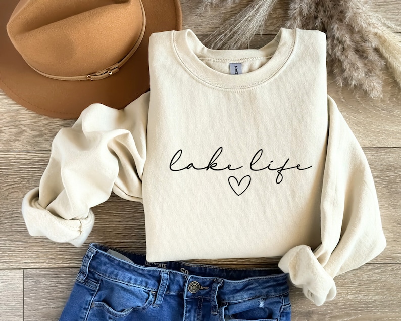 K&ouml;nnte beinhalten: Ein cremefarbener Pullover mit dem Text "lake life" und einem kleinen schwarzen Herz.