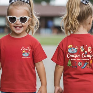 Pu&ograve; includere: Una t-shirt rossa a maniche corte con una piccola grafica sul petto che mostra una piscina, un ombrellone e il nome Emily sopra la scritta Cousin Camp 2026. Il retro presenta una grafica pi&ugrave; grande con la scritta Cousin Camp Mama and Papa 2026.