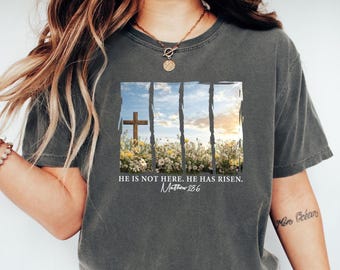 May include: Dark gray t-shirt featuring a graphic of a wooden cross in a field of flowers, framed by a window with a blue sky and clouds. The text reads "HE IS NOT HERE. HE HAS RISEN. Matthew 28:6."