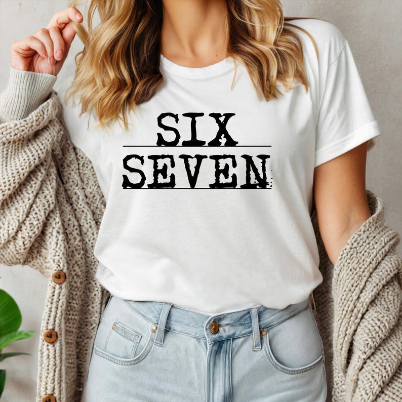 May include: White t-shirt with the words "SIX SEVEN" printed in black, with a line separating the words. The shirt is worn with blue jeans and a beige cardigan.