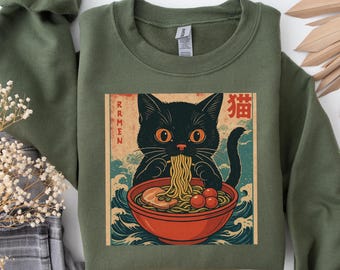 May include: Olive green sweatshirt featuring a graphic of a black cat eating ramen from a red bowl. The graphic includes the word "Ramen" in vertical text and Japanese characters. The cat has orange eyes and is surrounded by blue waves.