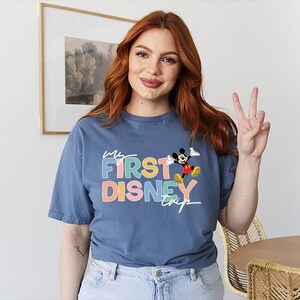 May include: A blue t-shirt with the text "My First Disney Trip" in colorful letters and a graphic of Mickey Mouse. The shirt is made of a soft material and has short sleeves.
