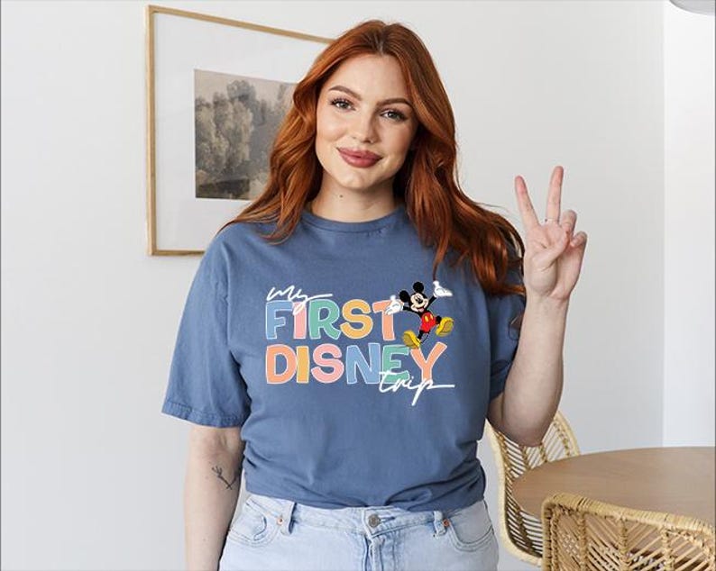 May include: A blue t-shirt with the text "My First Disney Trip" in colorful letters and a graphic of Mickey Mouse. The shirt is made of a soft material and has short sleeves.