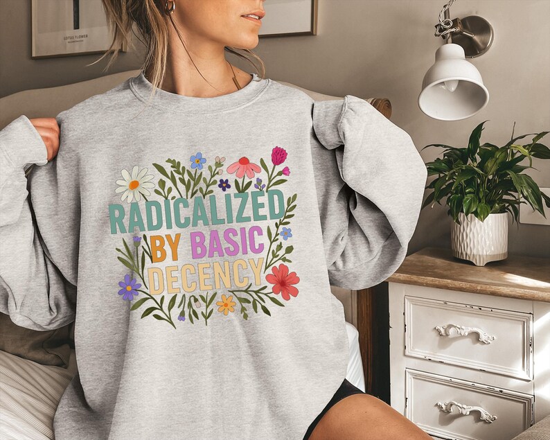 May include: A light gray sweatshirt with the text "RADICALIZED BY BASIC DECENCY" in colorful letters, surrounded by a floral wreath of daisies, pink, blue, and yellow flowers. The sweatshirt is long-sleeved.