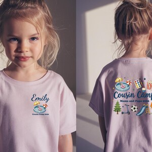 Pu&ograve; includere: Una t-shirt color viola chiaro con una piccola grafica sul davanti che riporta il nome Emily, una piscina e il testo Cousin Camp 2026. Il retro mostra una grafica pi&ugrave; grande con il testo Cousin Camp Mama and Papa 2026, inclusi icone di piscina, set di colori, popcorn, albero, monopattino, pallone da calcio, scivolo e ghiacciolo.