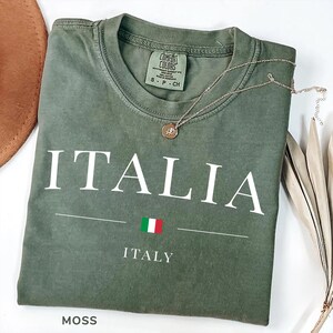 May include: A moss green t-shirt with the word "ITALIA" printed in white. Below the word is a horizontal line, the Italian flag, and the word "ITALY". A gold necklace with a pendant rests on the shirt.
