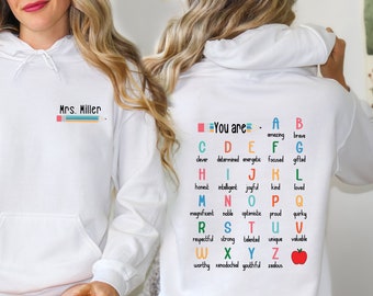 May include: White hooded sweatshirt with the text "Mrs. Miller" and a pencil graphic on the front. The back of the sweatshirt has the text "You are" and a colorful alphabet with positive words for each letter.