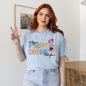 May include: Light blue t-shirt with the text "My First Disney Trip" in colorful block letters and a Minnie Mouse graphic. The shirt is made of a soft material and has short sleeves. The model is wearing light blue jeans.