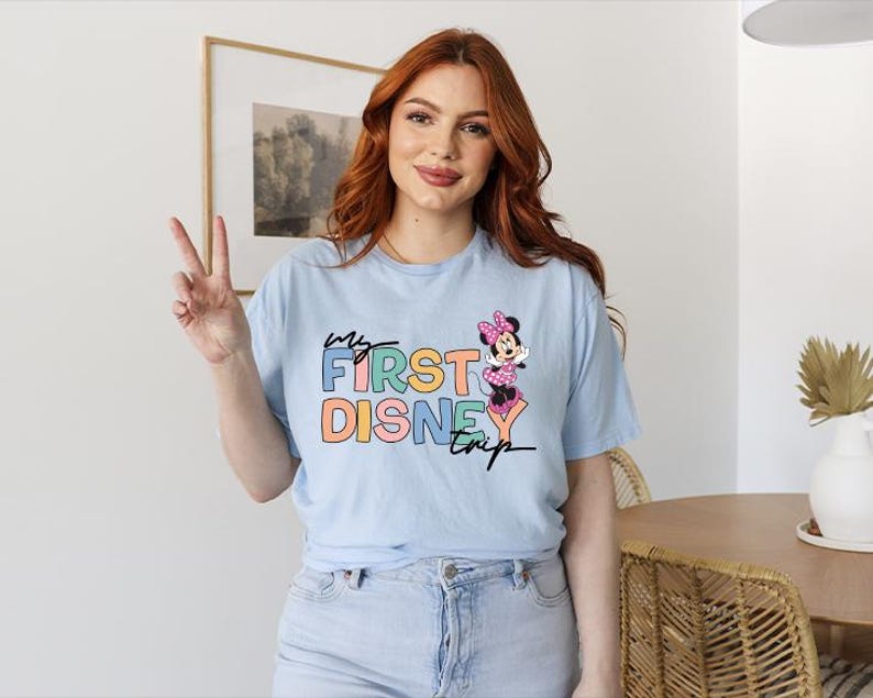 May include: Light blue t-shirt with the text "My First Disney Trip" in colorful block letters and a Minnie Mouse graphic. The shirt is made of a soft material and has short sleeves. The model is wearing light blue jeans.
