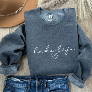 K&ouml;nnte beinhalten: Ein dunkelgrauer Pullover mit dem Schriftzug "lake life" in wei&szlig;er Schrift mit einem kleinen Herz darunter. Der Pullover ist gefaltet und auf einer Holzoberfl&auml;che mit einem braunen Hut und einer blauen Jeans gelegt.