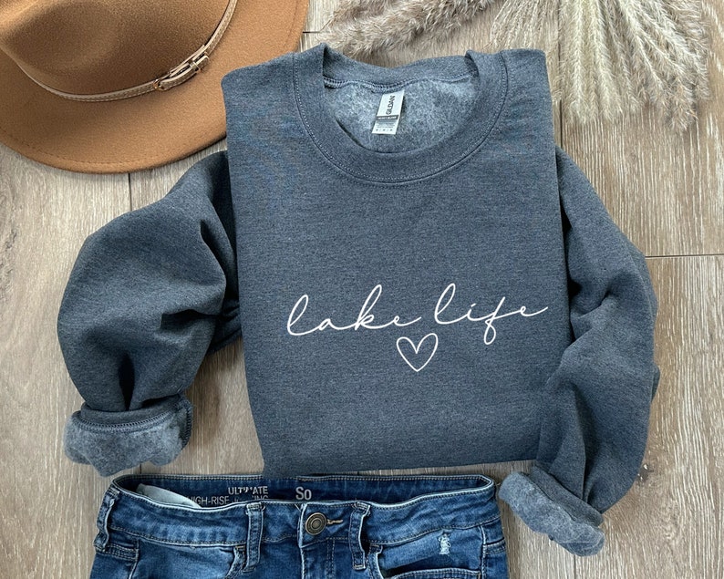 K&ouml;nnte beinhalten: Ein dunkelgrauer Pullover mit dem Schriftzug "lake life" in wei&szlig;er Schrift mit einem kleinen Herz darunter. Der Pullover ist gefaltet und auf einer Holzoberfl&auml;che mit einem braunen Hut und einer blauen Jeans gelegt.