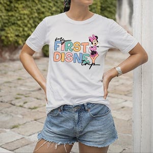 May include: White t-shirt with the text "My First Disney Trip" in colorful letters and a Minnie Mouse graphic. The shirt is paired with denim shorts. A silver bracelet is visible on the person's wrist.