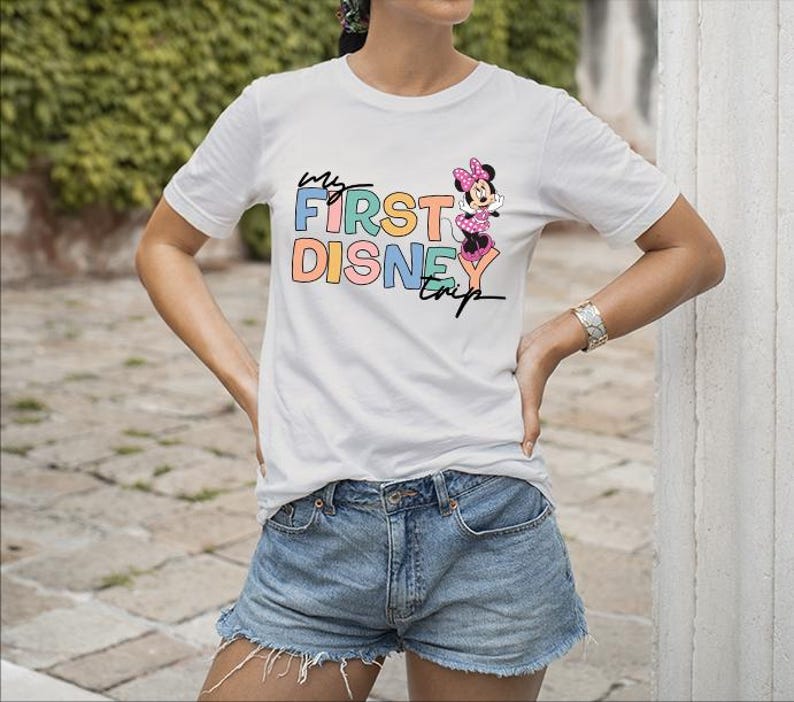 May include: White t-shirt with the text "My First Disney Trip" in colorful letters and a Minnie Mouse graphic. The shirt is paired with denim shorts. A silver bracelet is visible on the person's wrist.