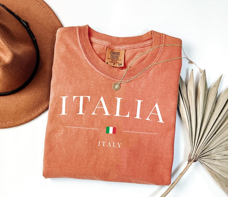 May include: A coral-colored t-shirt with the word "ITALIA" printed in white, along with the Italian flag and the word "ITALY". A gold necklace and a brown hat are also in the image.