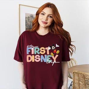 May include: Burgundy t-shirt with the text "My First Disney Trip" in colorful letters and a Mickey Mouse graphic. The shirt is a casual, oversized style.