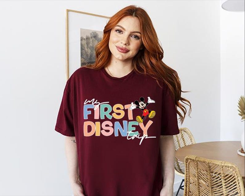 May include: Burgundy t-shirt with the text "My First Disney Trip" in colorful letters and a Mickey Mouse graphic. The shirt is a casual, oversized style.