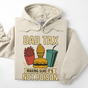 May include: Beige hoodie featuring the text "DAD TAX" and a graphic of fast food items: fries, ice cream, a burger, and a drink. The text "MAKING SURE IT'S NOT POISON" is below the graphic. The hoodie is folded on a white surface.