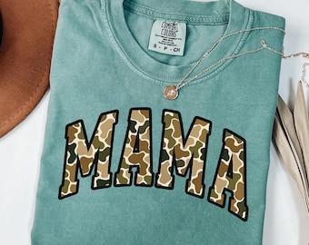 May include: A teal Comfort Colors t-shirt with the word "MAMA" in a camouflage pattern. A gold necklace with a pendant is on the shirt. The shirt is folded and placed on a white surface.