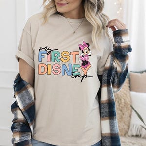 May include: Beige t-shirt with the text "my FIRST DISNEY trip" in colorful block letters and script. A Minnie Mouse graphic is on the right. The shirt is a casual style, perfect for a Disney vacation.