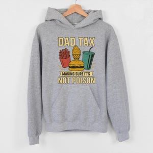 May include: A grey hoodie with a front pocket and a drawstring hood. The front features the text "DAD TAX" above a graphic of fries, ice cream, a burger, and a drink, with the text "MAKING SURE IT'S NOT POISON" below.