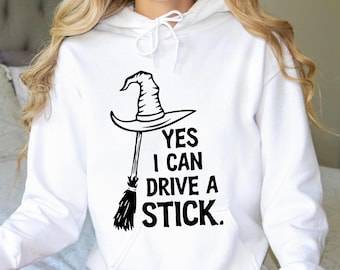May include: White hoodie with a black graphic of a witch's hat and broom. The text reads "YES I CAN DRIVE A STICK." The design is centered on the front of the sweatshirt.