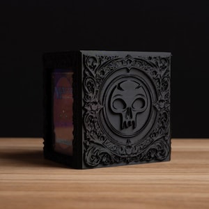 May include: A black, ornate box with a skull emblem on the front. The box has detailed carvings and a dark, gothic aesthetic. The side reveals a glimpse of the contents, suggesting a card game theme.