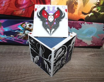 May include: A white cube-shaped box with a black and white design on the sides. The top of the box is white and features a black and red design with a woman's face in the center. The box is made of cardboard and has a blue interior.