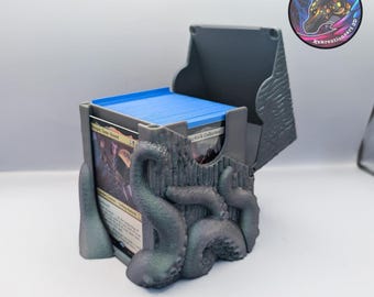 May include: A gray 3D-printed card storage box with an open lid, featuring a textured design with tentacles. The box holds a stack of blue-edged cards. A circular sticker with a horse-like creature is visible on the right.