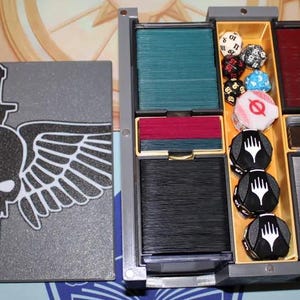 May include: A gray box with a skull and wings design on the lid. The box has multiple compartments for storing cards and dice. The compartments are filled with various colored cards and dice.