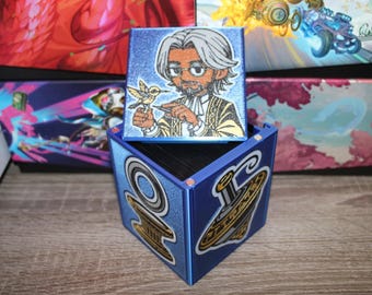 May include: A blue cube-shaped box with a hinged lid. The lid features a colorful illustration of a man with a beard holding a small bird. The box has a blue and gold design on the sides.