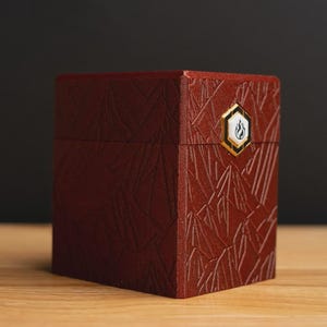 May include: A maroon-colored, square box with a textured, geometric pattern. The box features a gold-trimmed emblem with a flame design. The box is set on a wooden surface against a dark background.