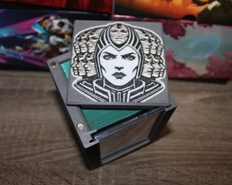 May include: A black and silver card deck box with a graphic of a woman's face with multiple faces surrounding her. The box is made of plastic and has a hinged lid.