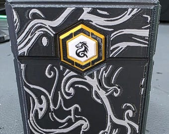 May include: Black rectangular box with silver swirling patterns and a gold and white emblem featuring a dragon. The box has a textured surface and appears to be 3D printed. The emblem is a hexagon shape.