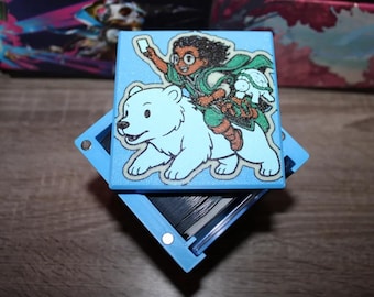 May include: A blue box with a hinged lid. The lid features a colorful illustration of a person riding a polar bear. The person is holding a book and wearing a green cloak. The box is open and appears to be a storage box for cards.