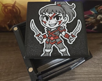 May include: Black card storage box with a cartoon warrior graphic on the lid. The warrior is depicted with a ponytail, holding two swords, and wearing red and gray armor. The box is open, revealing a stack of cards.